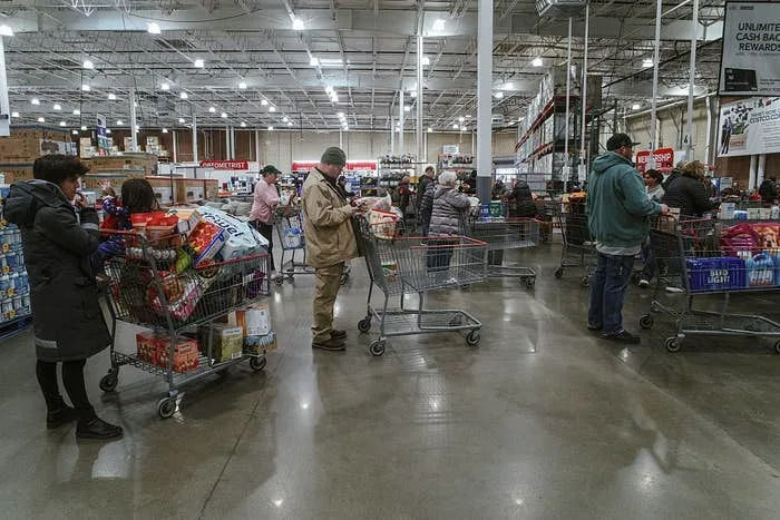 Customers with full carts line up in a busy warehouse store, waiting at the checkout