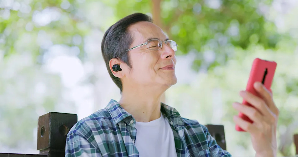 A person with glasses smiles while holding a smartphone outdoors, appearing to enjoy music through wireless earbuds