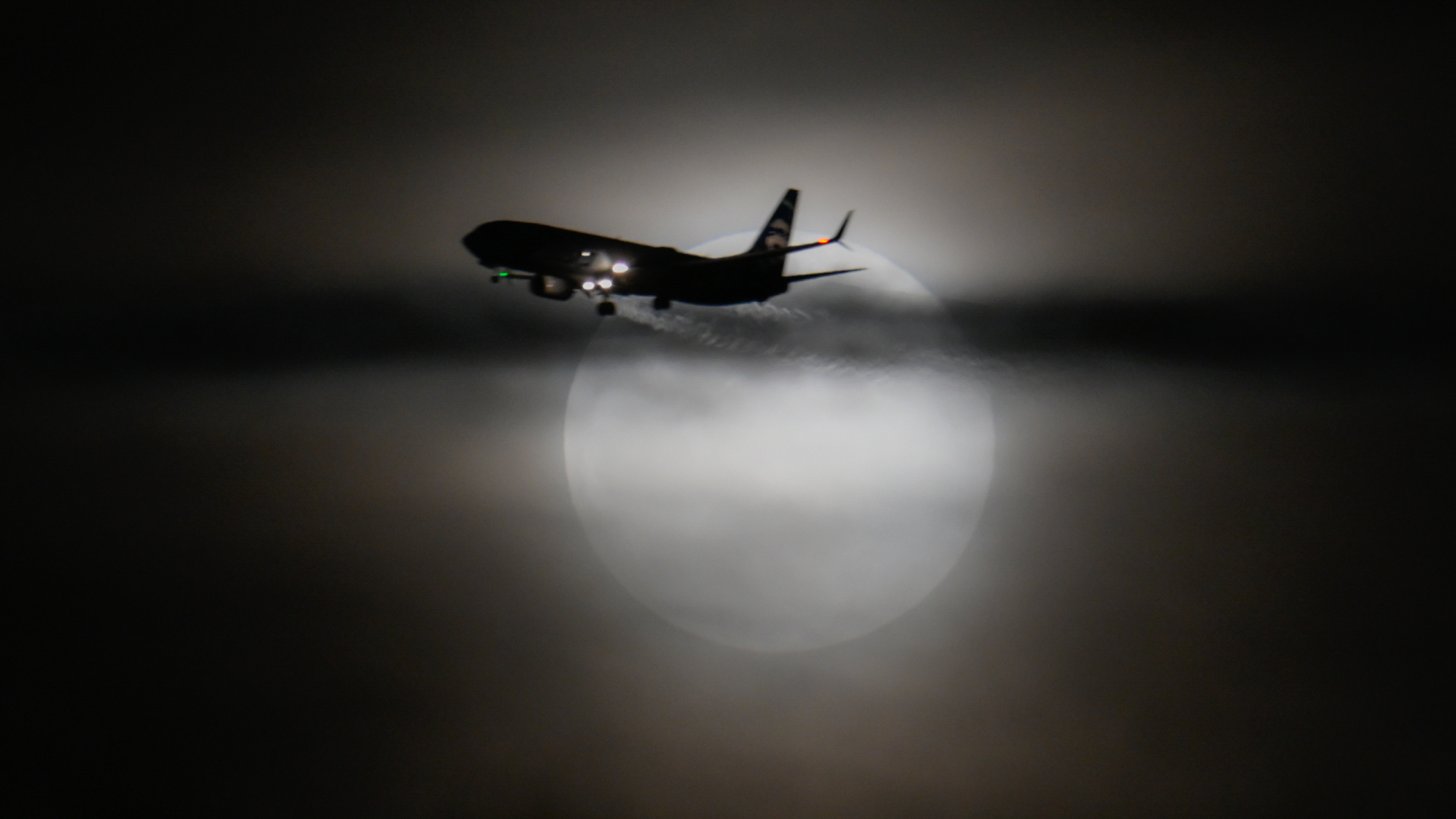 The silhouette of a passenger jet is pictured crossing a hazy full moon against a dark sky.