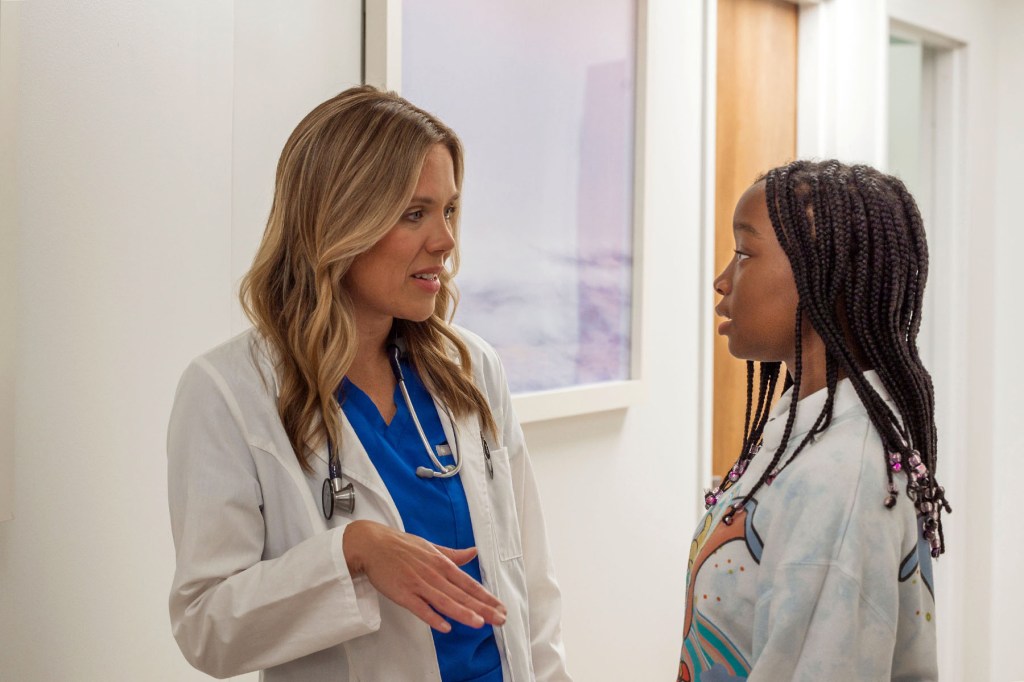 Dr. Annie Andrews, wearing a white coat and stethoscope, speaks with a young girl in a medical office hallway.