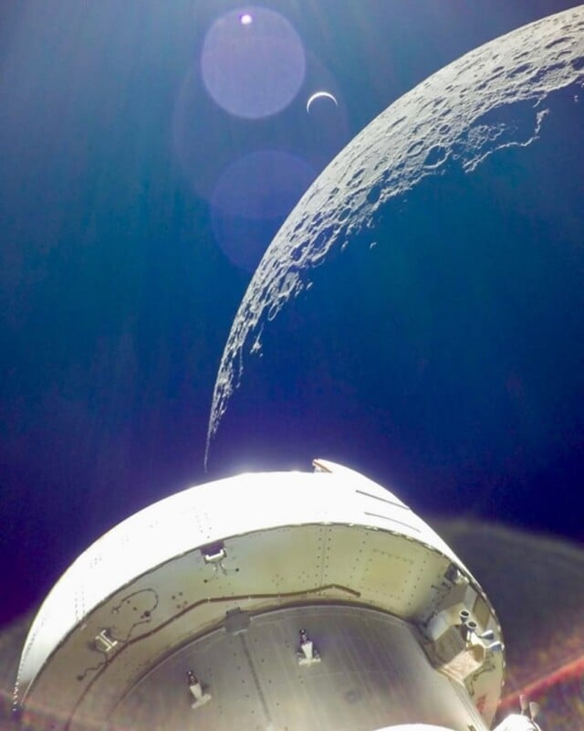A spacecraft orbits near the Moon’s rugged, sunlit surface with Earth visible as a crescent in the distant blackness of space. Lens flares are visible near the top of the image.
