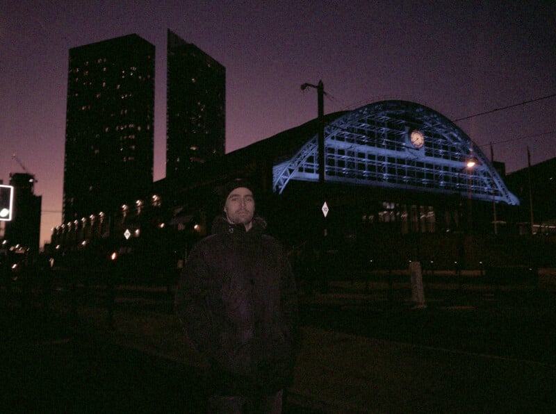 A person in a dark coat and hat stands in front of a large, glass-arched building illuminated with blue light at dusk, with tall buildings in the background against a purple sky.