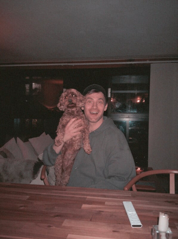 A smiling person in a gray hoodie and cap sits at a wooden table, holding a small brown dog. A remote control is on the table, and soft lighting illuminates a cozy indoor setting in the background.