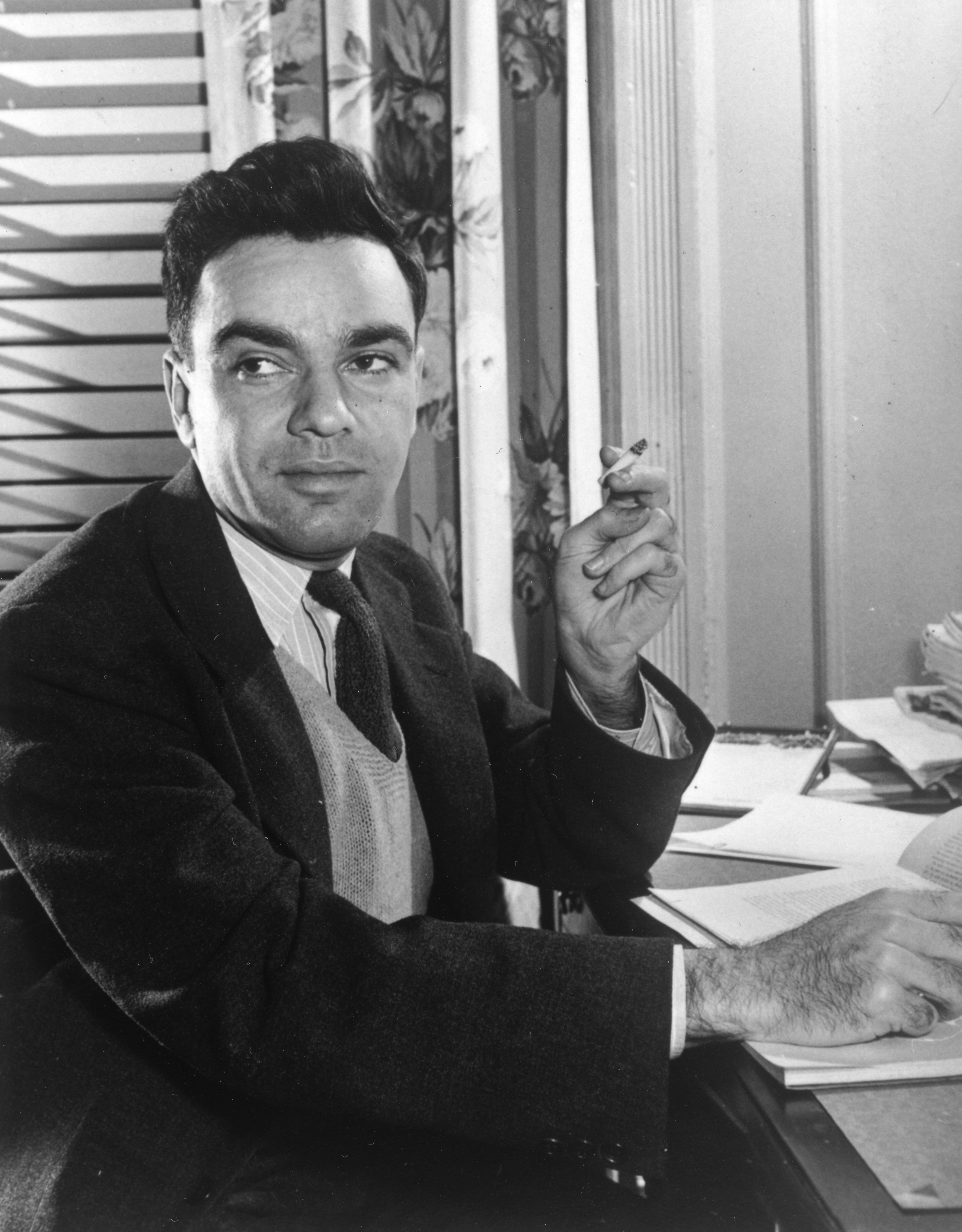 A black and white photo shows a man with dark hair and dark eyes, wearing a suit, vest and tie, sitting at a desk full of papers with a half burnt cigarette in his left hand. He looks to the left of the camera