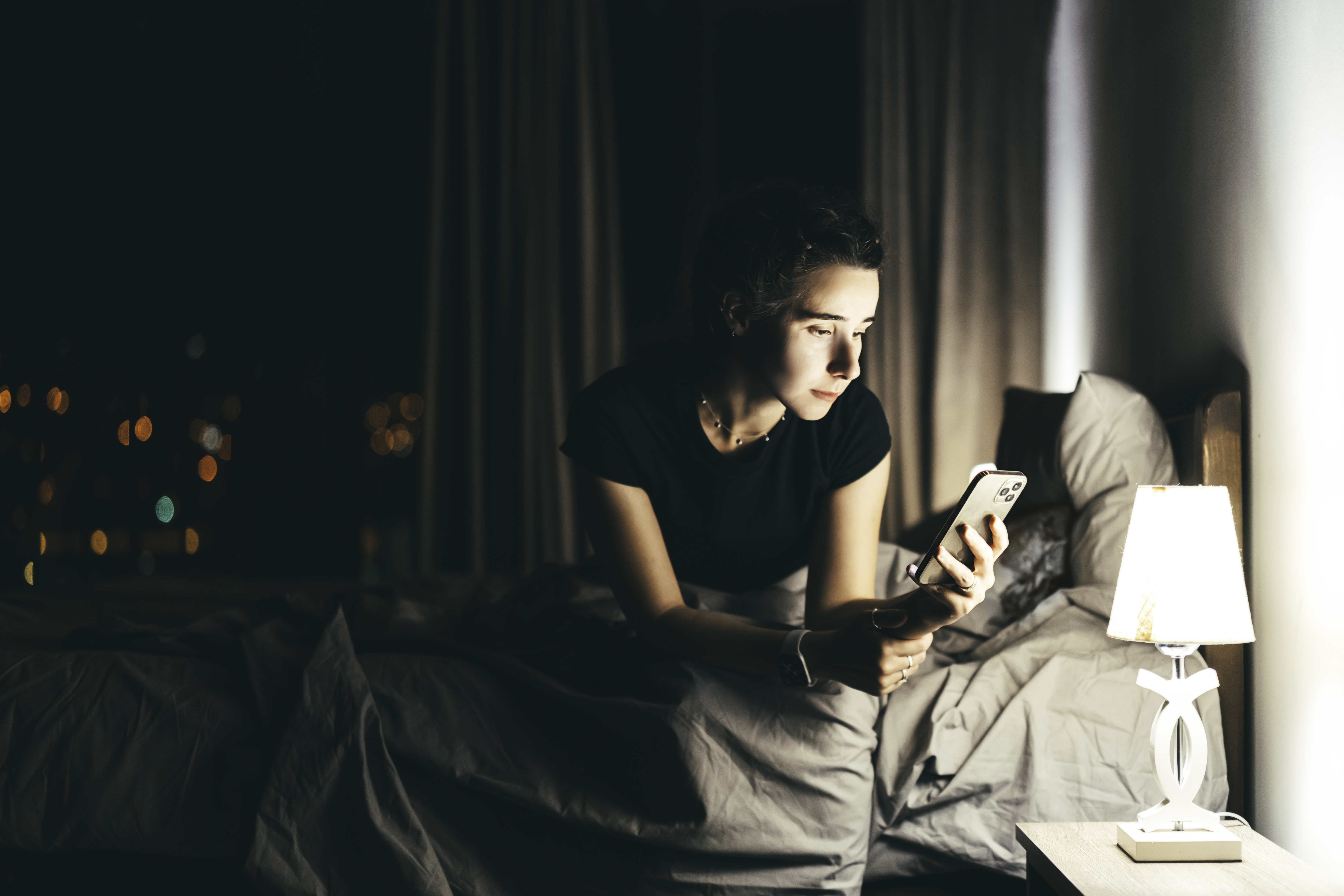 A woman sitting in her bed at night, looking at her phone. She is illuminated by the light from her bedside lamp.
