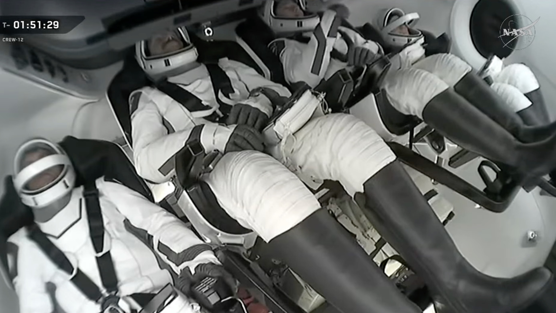 four astronauts in white and black spacesuits sit inside a space capsule on earth