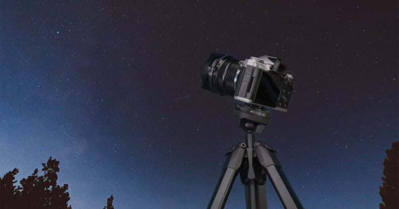 A digital camera mounted on a tripod is set up outdoors at night, aimed at a clear, star-filled sky with silhouettes of treetops visible at the bottom of the image.