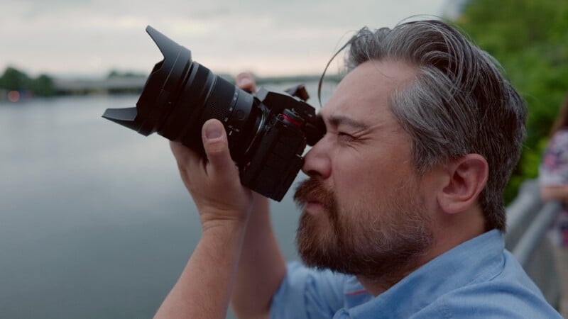 A man with a beard and gray-streaked hair, wearing a light blue shirt, holds a camera up to his eye and takes a photo outdoors near a body of water, with trees and a cloudy sky in the background.