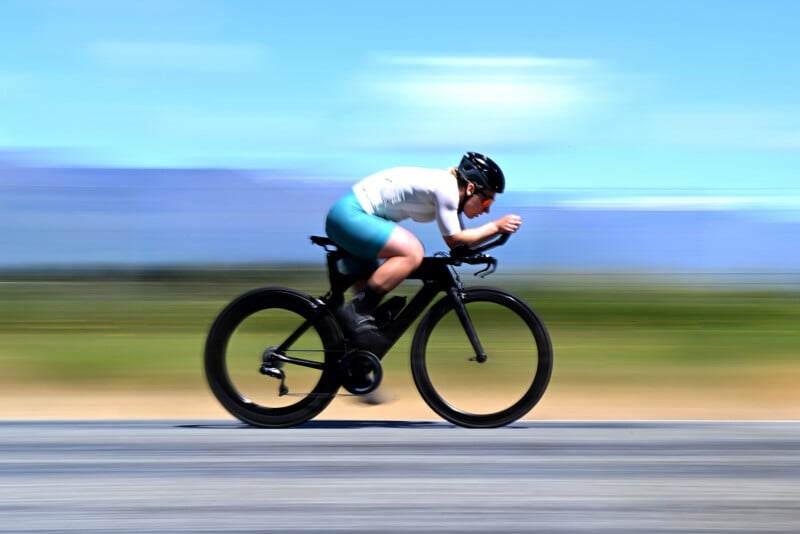 A cyclist wearing a helmet and teal shorts rides a racing bicycle at high speed on a road, with a blurred background indicating motion under a clear blue sky.