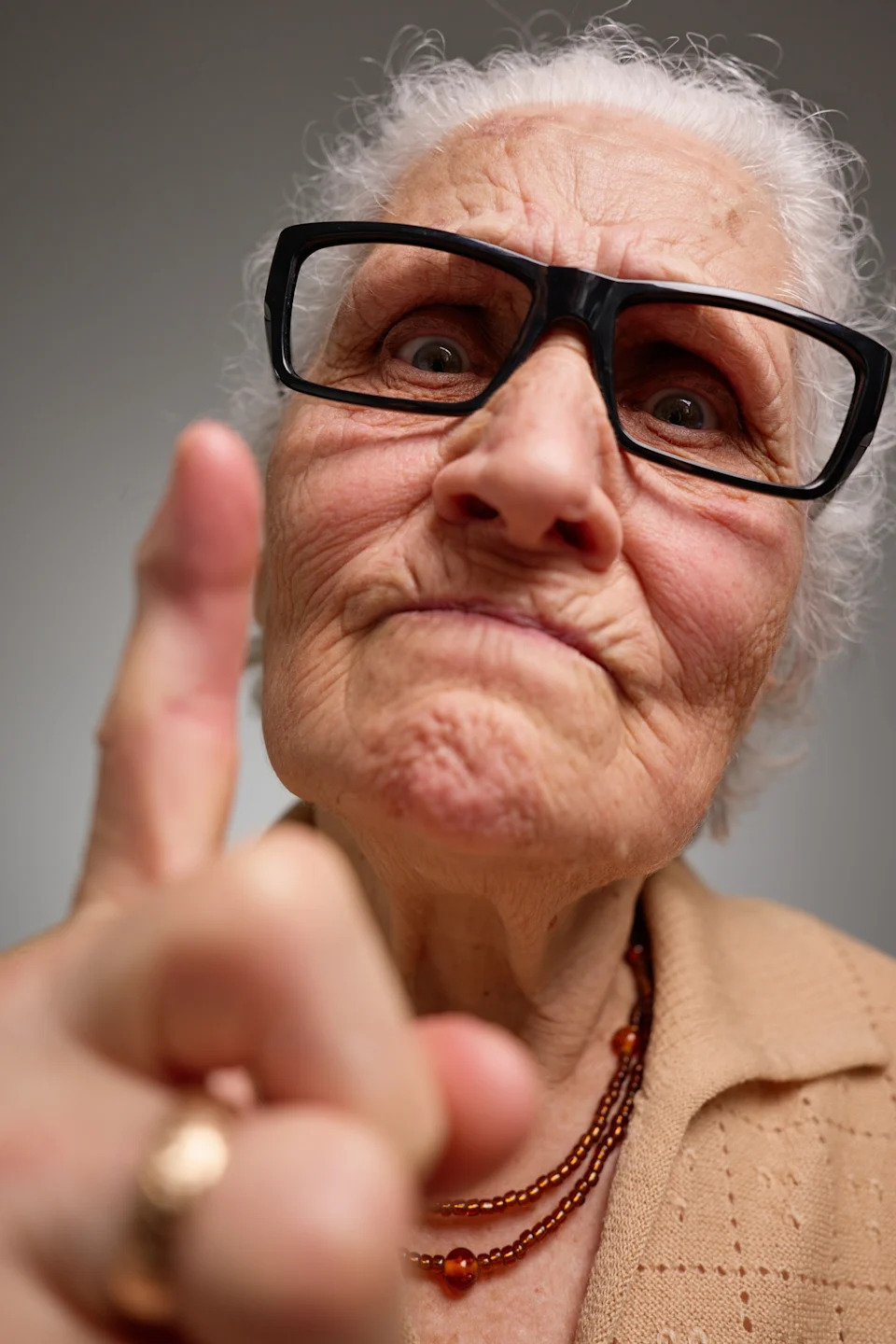 Elderly woman with glasses points a finger toward the camera, wearing a bead necklace and textured cardigan, with a focused expression