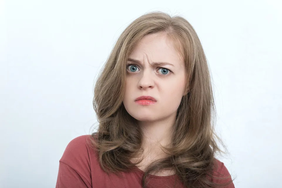 Person with wavy hair and a puzzled, skeptical facial expression, wearing a casual top. Neutral background