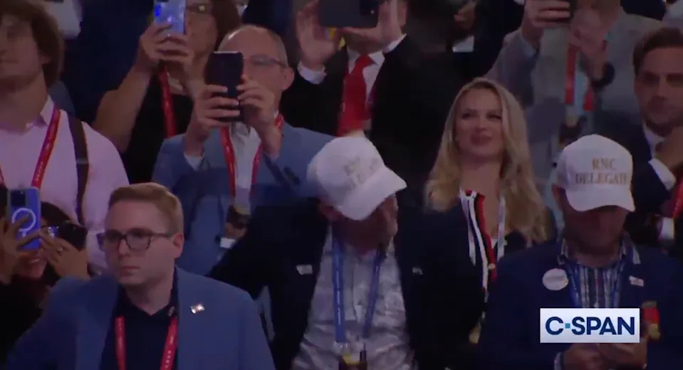 People at an event, many taking photos with phones. Some are wearing hats, and one person is filming