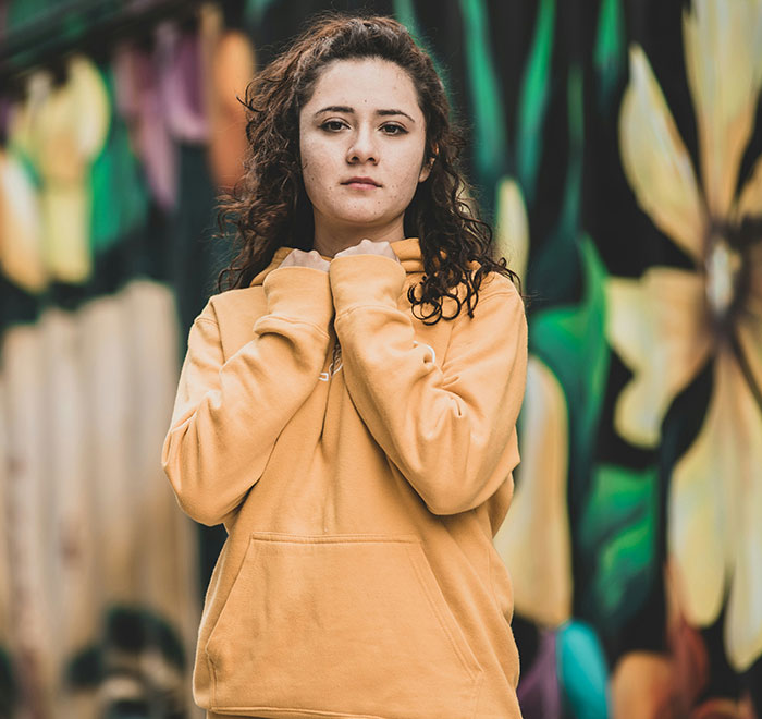 Young woman in a yellow hoodie posing thoughtfully in front of a colorful mural, adding a weirdly hilarious vibe to the feed.