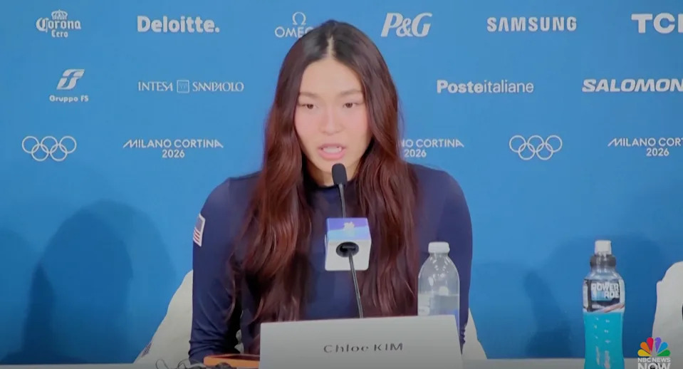 Person speaking at a press conference, seated at a table with microphones and drinks, Olympic logos and sponsor names on the background