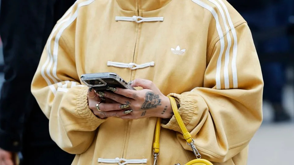 A guest wears dark sunglasses, a mustard yellow suede Adidas Tang jacket with white toggle closures, oversized beige cotton trousers, and a bright yellow Vivienne Westwood heart-shaped leather handbag, outside Jeanne Friot, during the Menswear Fall/Winter 2026-2027 as part of Paris Fashion Week on January 20, 2026 in Paris, France. - Claudio Lavenia/Getty Images