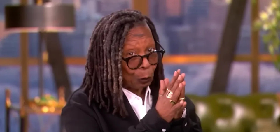 A person with glasses and long hair, wearing a suit with rings, gestures with hands clasped on a TV talk show set