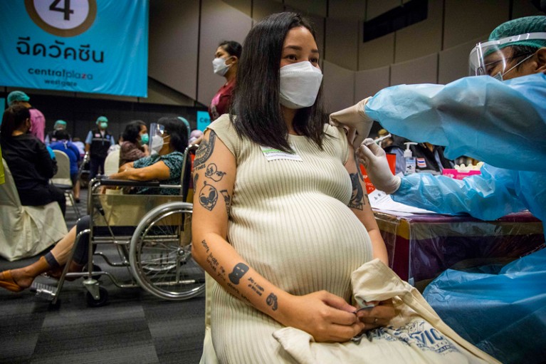 In a large busy room, a pregnant woman wearing a face mask receives a vaccine from a health worker wearing PPE.