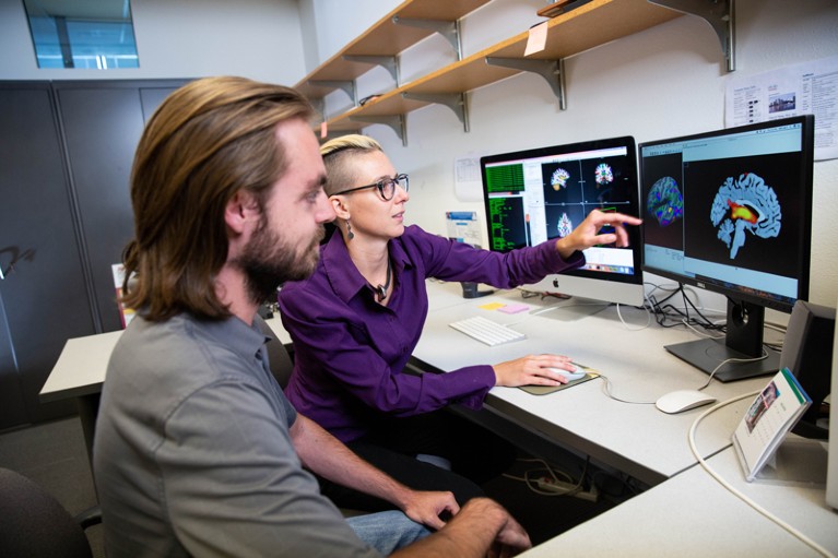 Two people sit at a desk, looking at computer screens displaying brain-scans images.