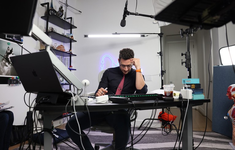 A behind the scenes view of Doctor Mike reading at his desk surrounded lights, props and microphones during a break in recording.