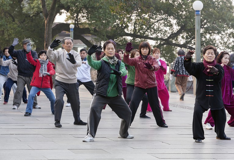 A group of senior citizens in warm clothing exercise, standing with arms raised, in a park.