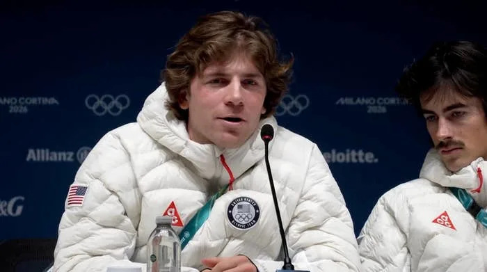 Person speaking at a press event with Olympic logos in the background, dressed in a puffy white jacket