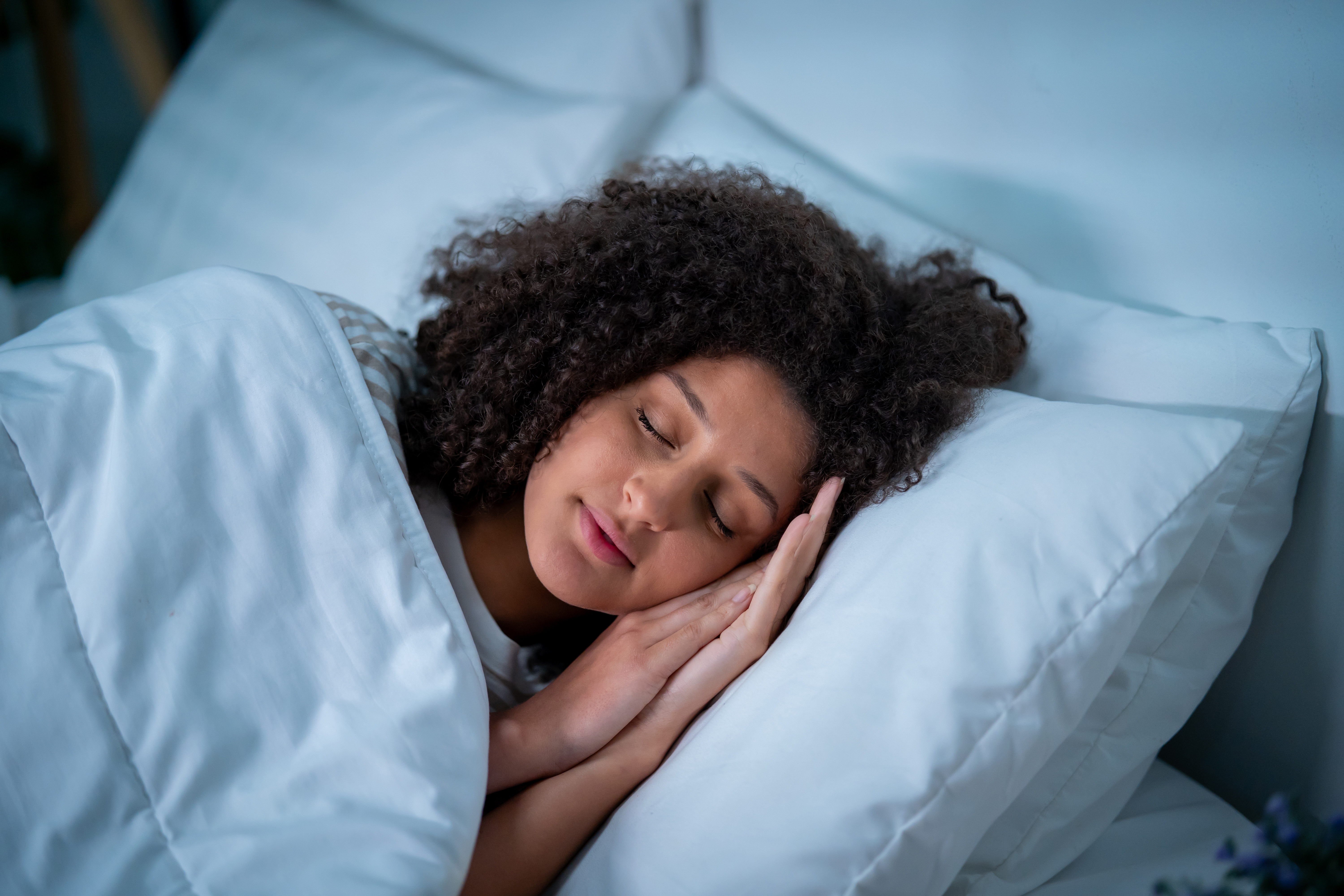 A woman lies asleep on her side in bed with her hands placed together next to her face.