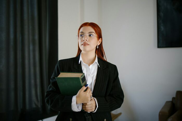 Young woman with red hair and glasses holding a green book, appearing thoughtful while standing indoors, expressing daily worries.