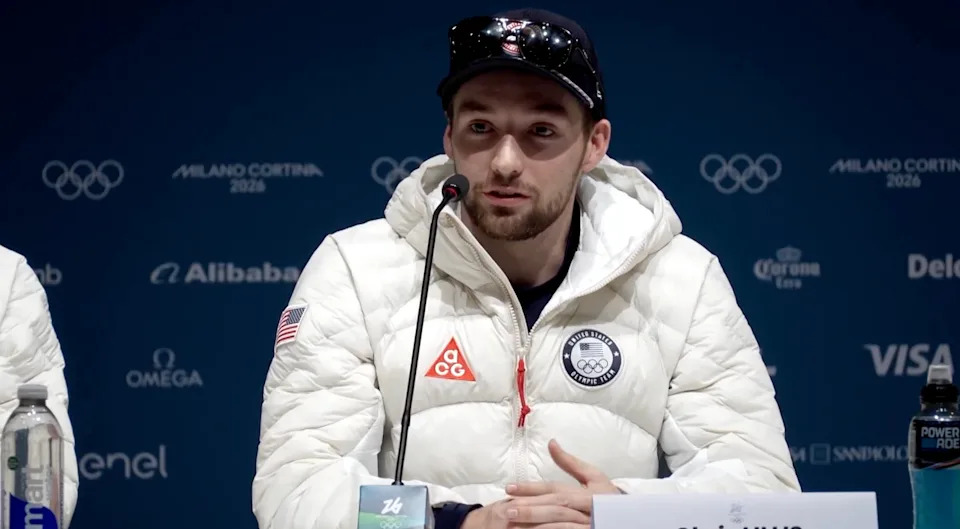 Athlete at press conference in a branded jacket with logos speaks into a microphone, wearing a cap and sunglasses, against a backdrop of sponsor logos
