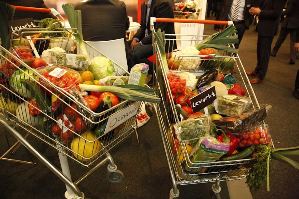 Two shopping carts filled with a variety of fresh produce, including leeks, lettuce, peppers, and packaged items, seen in a busy indoor market setting
