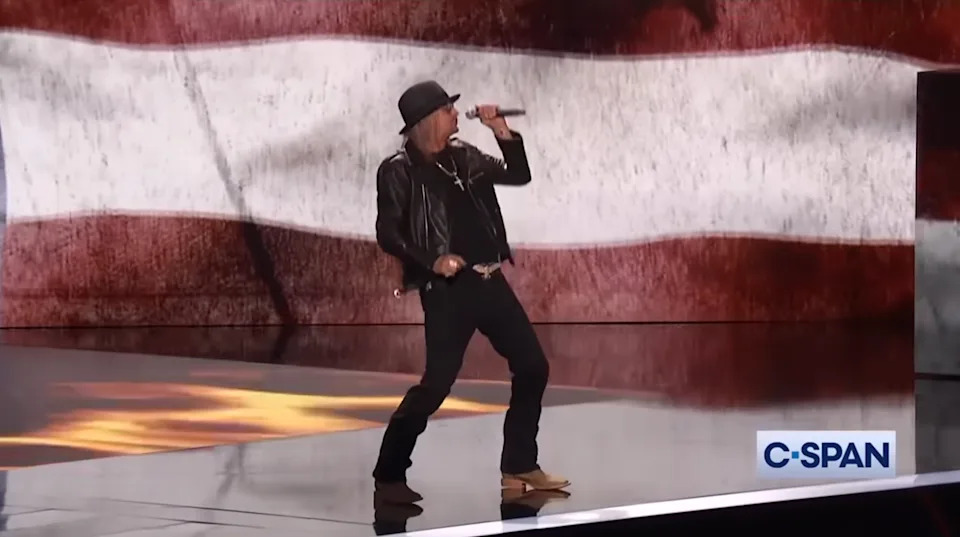 Performer wearing a black leather jacket and hat singing on stage with a large flag backdrop, C-SPAN logo in the corner