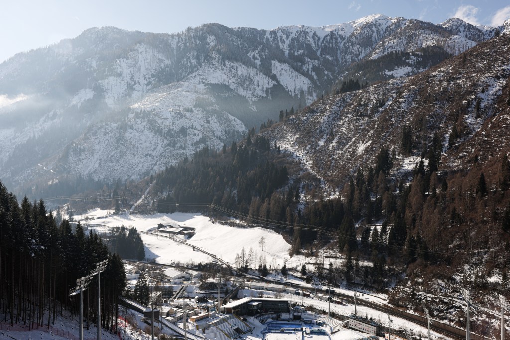 General view of Predazzo Ski Jumping Stadium for the Milano Cortina 2026 Winter Olympics, with snow-covered mountains and trees in the background.