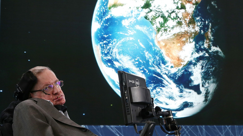 Stephen Hawking sits in his wheelchair before a projection of Earth from space.