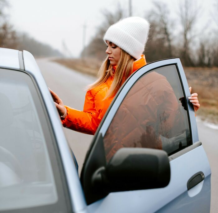 Woman in orange jacket and white beanie opening car door on a quiet road, reflecting women's daily worries and peaceful bliss contrast.