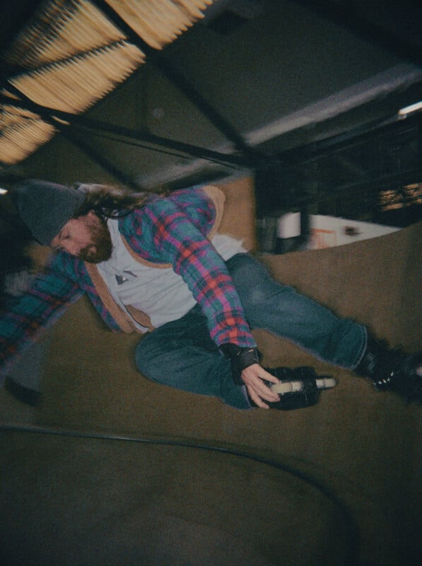 A person wearing a beanie, flannel shirt, and jeans is rollerblading on an indoor skate ramp, captured mid-air in motion blur. The setting appears to be an industrial skate park.