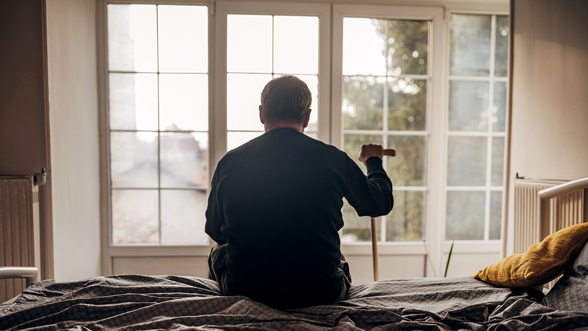 old man sits on bed looking out window