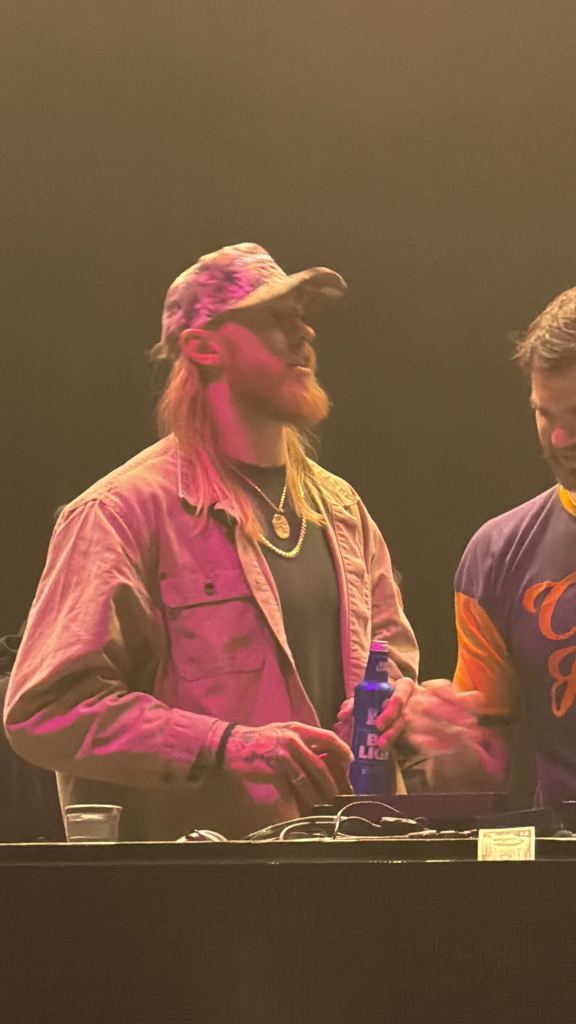George Kittle, with long hair and a beard, in a hat, gold chains, and a tan jacket, is seen next to another person with a bottle of Bud Light.