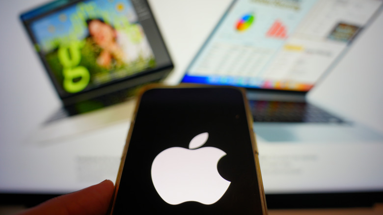 Picture of an iPhone with the Apple logo on the screen and pictures of MacBooks in the background