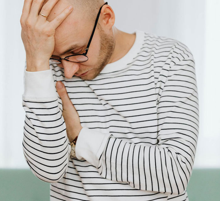Man wearing glasses and striped shirt looking stressed, holding his head and crossing his arms in an indoor setting.
