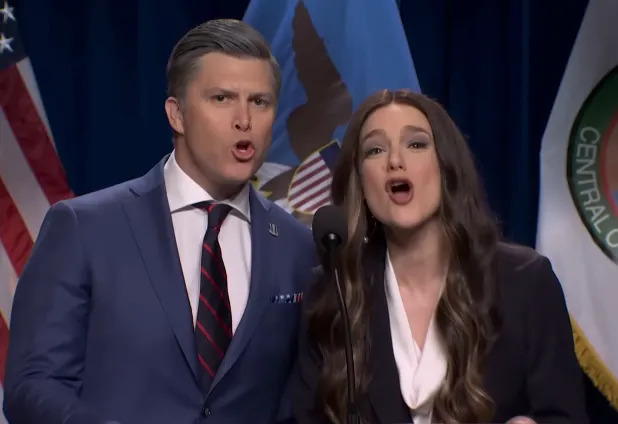 Two individuals on stage singing or speaking enthusiastically into a shared microphone, both dressed in formal attire with American flags in the background