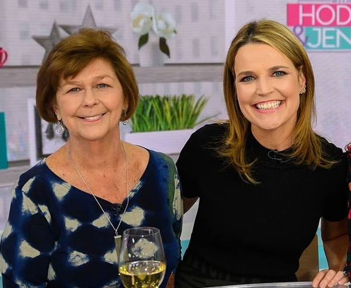 Two women, one wearing a floral-patterned top and the other in a solid black top, are standing next to each other and smiling, likely for a photo shoot in a studio.
