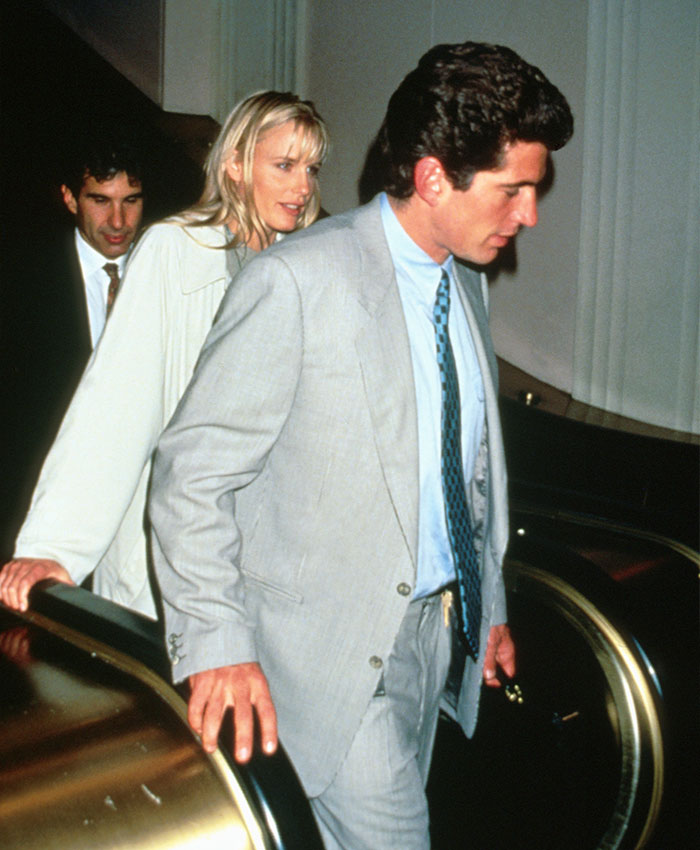 Daryl Hannah with JFK Jr. and another man, dressed formally, walking down an escalator in a public setting.