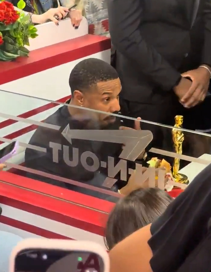 Michael B Jordan sitting behind a glass counter with an Oscar statue, grabbing food after the Oscars event.