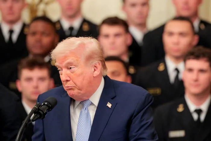 A man in a suit speaking at a podium, with a group of people in uniformed attire in the background