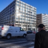 A concrete and glass building is seen across the street as an older man waits at a busy crosswalk.