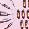 Clear syringes face towards a line of clear vials. Light pink background. 