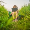 Happy child sitting on the field holding tablet. Boy sitting on the grass on sunny day. Home schooling or playing a tablet.