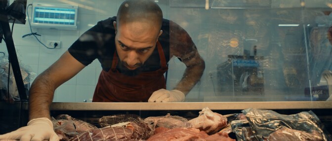 A bald man with medium skin tone leans over meat at a butcher counter. He's wearing white gloves and an apron. 