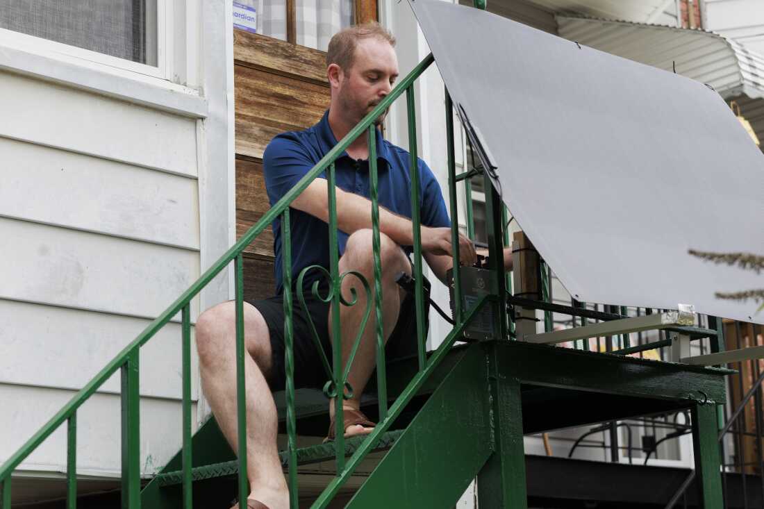 Sitting outdoors on his back steps, which are metal and painted green, Craig Keenan installs a plug-in solar panel in front of the railing at the top.