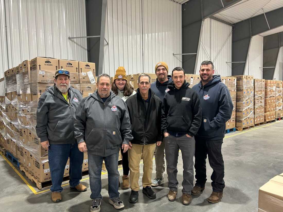 Mike Puglisi and six of his family members stand inside a storage facility in which cardboard boxes of eggs are stacked tall on pallets.