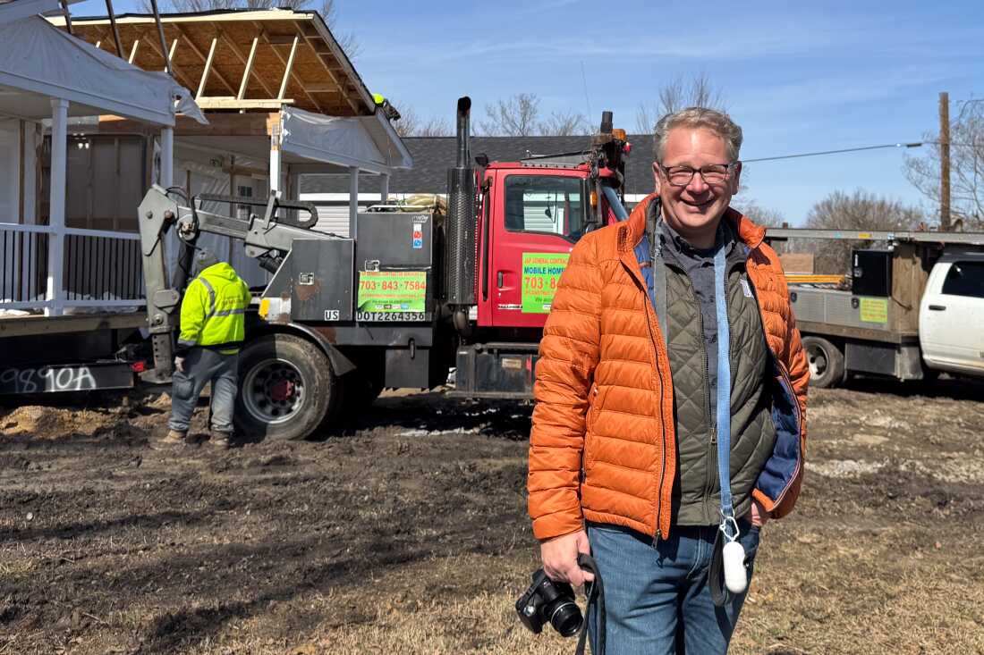 Developer Tom Heinemann stands at a site in Petersburg, Virginia, where the two halves of a four-bedroom factory-built home are getting pushed together in the background. A truck and worker are also in the background.